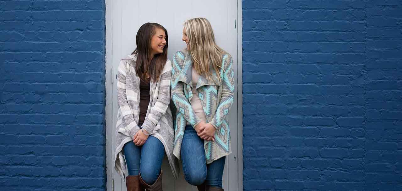 Zwei Freundinnen stehen in einer Türöffnung vor einer blauen Wand und lächeln sich an.